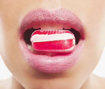 A woman holds glossy red and white striped candy in her teeth. Implicating that the person will use the mint for fresher breath.